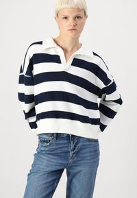 Pullover listrado em branco e azul-marinho com decote em V e gola, apresentando ombros caídos e um corte descontraído, combinado com jeans azuis.