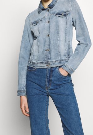 Person iført en lyseblå denimjakke med knapper og blå jeans med høj talje, den ene hånd i lommen og den anden afslappet ned langs siden.