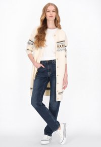 Krém színű kardigán mintás ujjakkal, kék farmerral és fehér tornacipővel párosítva. A kardigán arany gombokkal és puha textúrával rendelkezik.