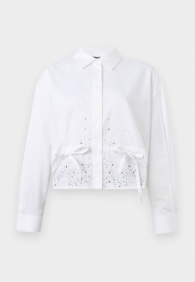 Chemise blanche à manches longues, courte, avec col, boutons sur le devant, ornements en strass et deux nœuds attachés près de la taille sur un fond uni.