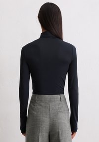 Frau mit langen dunklen Haaren, die einen schwarzen, langärmligen Rollkragenpullover trägt, in die hoch taillierte graue Stoffhose gesteckt, steht vor einer weißen Wand.