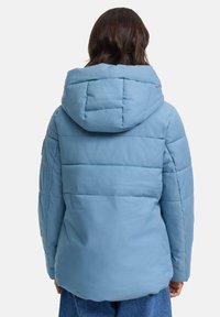 Person in einer hellblauen gepolsterten Jacke mit Kapuze, von hinten gezeigt, und blauen Jeans vor einem schlichten Hintergrund.
