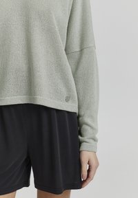 Lichtgroene gebreide trui met lange mouwen, voorzien van een ronde onderkant en een kleine metalen accent, gecombineerd met zwarte shorts.