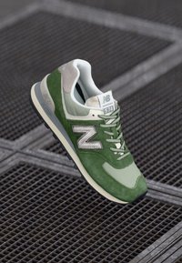 Žalios ir pilkos New Balance 574 sportbačiai su balta logo ir raišteliais, padėti ant metalinės grotelės paviršiaus.