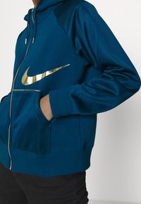 Person som bär en blå zip-up hoodie med en glänsande guld Nike-swoosh-logga, händerna i framfickorna, i kombination med svarta byxor.