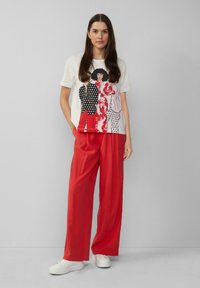 T-shirt blanche à manches courtes avec un imprimé graphique de trois figures vêtues de motifs, associée à un pantalon large rouge et des baskets blanches.