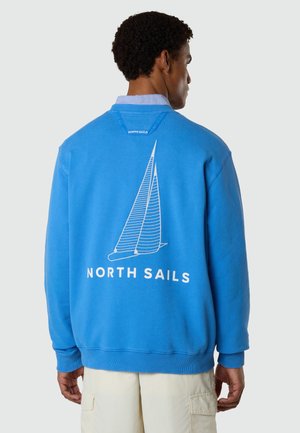 Homme portant un sweat-shirt bleu avec un motif de voilier blanc et le texte "NORTH SAILS" dans le dos, associé à un short cargo beige.