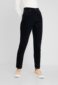 Pantalon en velours côtelé noir avec taille haute, présentant une coupe droite, des poches avant et des détails en métal sur les boutons. Texture lisse.