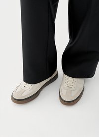 Baskets blanches en cuir lisse avec des accents en daim, dotées d'un design classique à lacets et d'une semelle en caoutchouc marron. Pantalon noir partiellement visible.