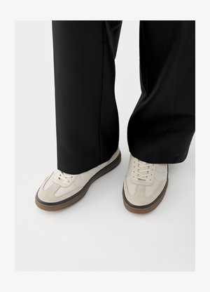 Weiße Sneaker aus glattem Leder mit Wildlederakzenten, ausgestattet mit einem klassischen Schnürdesign und einer braunen Gummisohle. Schwarze Hose teilweise sichtbar.