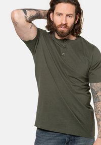 Camiseta henley de manga corta en verde oliva con detalle de tres botones. Tejido suave y texturizado con un ajuste relajado, combinada con vaqueros azules.