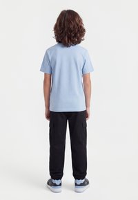 Dítě s kudrnatými hnědými vlasy, oblečené v světle modrém tričku s krátkými rukávy, černých kalhotách typu cargo a šedých teniskách, stojící zády.