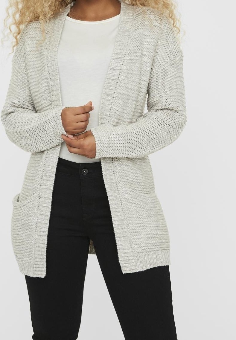 Cardigan gris clair en maille avec devant ouvert, détails en côtes et deux poches avant. Porté sur une chemise blanche et un pantalon noir.