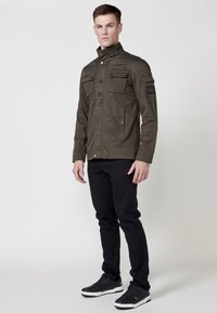 Jachetă verde oliv cu guler înalt, două buzunare pe piept și buzunare laterale. Purtată peste pantaloni negri slim-fit și adidași negri.