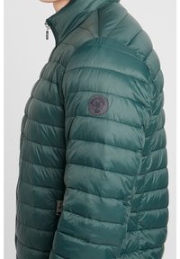 Grøn pufferjakke med høj krave, vandret quiltet design og et rundt logo-patch på venstre skulder. Lynlåslomme i siden.
