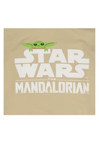 T-shirt beige con grande testo bianco "Star Wars" e logo di "The Mandalorian", con un grafico di un personaggio verde con grandi orecchie sopra.