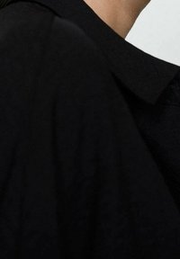 Vêtement noir à col avec un tissu texturé, présentant une coupe ample et un drapé subtil. Le col est structuré et soigneusement fini.