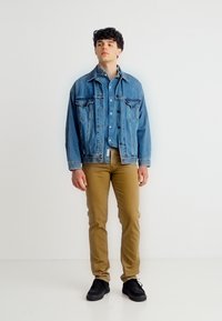 Νεαρός άνδρας όρθιος, φορώντας στρωμένη μπλε τζιν μπουφάν και πουκάμισο, καφέ παντελόνι και μαύρα παπούτσια μπροστά σε απλό λευκό φόντο.