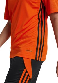 Camiseta deportiva naranja de manga corta, con tres rayas negras a lo largo del costado y la marca "AEROREADY" cerca del dobladillo. Pantalones cortos deportivos negros que complementan.