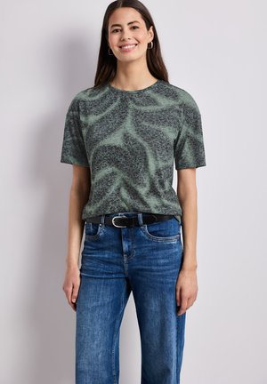 Femme aux longs cheveux foncés portant un t-shirt gris et noir à motifs, rentré dans un jean bleu avec une ceinture noire, souriante sur un fond uni.