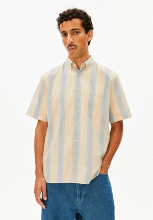 Mann mit kurzen lockigen Haaren und Schnurrbart, der ein beige und hellblau vertikal gestreiftes kurzärmeliges Hemd und blaue Jeans trägt.
