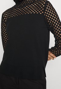 Haut noir à manches longues avec corps uni et superposition à grands trous découpés, col montant et ourlet côtelé.