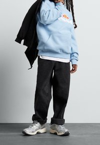 Sweat à capuche bleu clair avec logo blanc, jean noir surdimensionné et baskets argentées avec semelles crème. La personne porte une veste bomber noire.