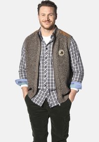 Herren brauner Strickwesten mit Lederakzenten, getragen über einem blau-weißen karierten Hemd. Die Weste verfügt über ein Logo-Patch und einen Reißverschluss vorne.