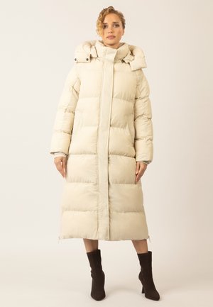 Light beige long puffer coat with a high collar, snap closures, and a quilted design. Worn with dark, fitted ankle boots.
