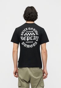 Άνδρας με κυματιστά σκούρα μαλλιά που φοράει μαύρο μπλουζάκι με λευκό κείμενο και καρό γραφικό στην πλάτη, συνδυασμένο με ανοιχτόχρωμο καφέ παντελόνι cargo.