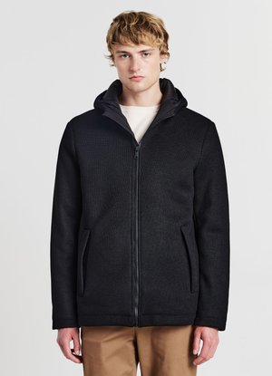 Junger Mann mit blondem Haar, der eine schwarze Jacke mit Reißverschluss und Kapuze über einem weißen Hemd und braunen Hosen trägt, vor einem schlichten Hintergrund stehend.