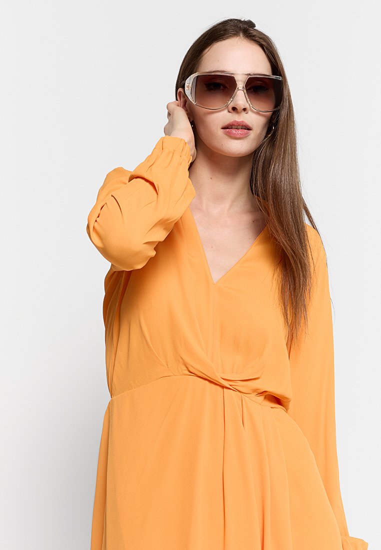 Kobieta w dużych brązowych okularach przeciwsłonecznych i pomarańczowej sukience z długim rękawem o dekolcie w kształcie V, dotykająca włosów w pobliżu prawego ucha na tle białym.