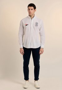 Camisa blanca de botones con logotipos bordados en el pecho, combinada con pantalones oscuros de botones y zapatillas blancas. Tejido suave y ajuste entallado.