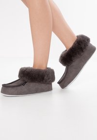 Pantofole in suede grigio con un morbido risvolto in pelliccia grigia scura, design piatto, punta arrotondata e suola bianca morbida. Nessun hardware o decorazione visibile.