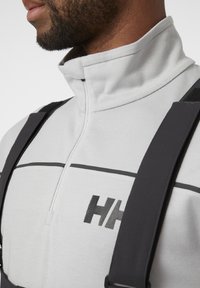 Sudadera de forro polar gris claro con cuello alto, cremallera negra y logotipo negro. Presenta correas negras ajustables para mayor soporte. Textura suave.
