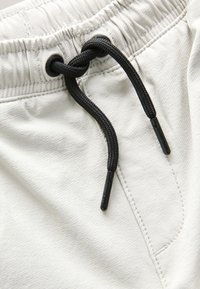 Primo piano di pantaloni in tessuto beige chiaro con vita elasticizzata e coulisse nera annodata vicino agli occhielli in metallo.