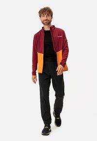 Leichte Jacke mit Reißverschluss in Rot und Orange mit Netzdetails, kombiniert mit schwarzen dehnbaren Hosen und schwarzen Turnschuhen. Hat eine lockere Passform.