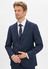 Granatowy garnitur z tkaniny o teksturze, zapinany na jeden guzik, z klapami w szpic i kieszeniami bocznymi; w zestawie z białą koszulą i wąskim krawatem w paski.