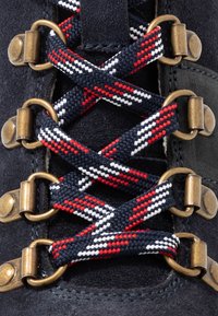 Bota de ante azul marino con ojales de bronce, que presenta un diseño de cordones trenzados en rojo, blanco y azul marino. Primer plano del detalle del cordón.