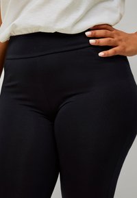 Kvinde iført højhalset sorte leggings med en hvid top, hånd hvilende på hoften, negle malet lyserøde, på grå baggrund.