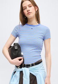 Νεαρή γυναίκα που φοράει εφαρμοστή μπλούζα με μπλε και λευκές ρίγες, κρατώντας μια μαύρη τσάντα, με ανοιχτό μπλε μπουφάν δεμένο γύρω από τη μέση, σε λευκό φόντο.