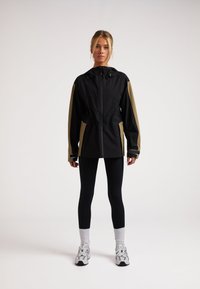 Svart och olivgrön vindjacka med huva, blixtlås, sidofickor och elastiska muddar, kombinerad med svarta leggings och sneakers.
