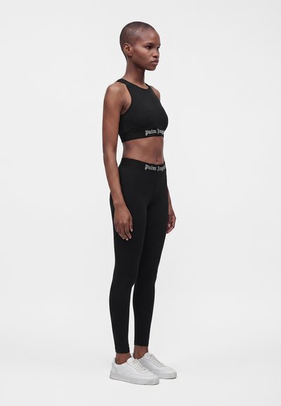 Γυναίκα που στέκεται με μαύρο crop top και κολάν με το λογότυπο "Palm Angels", φοράει λευκά αθλητικά παπούτσια, σε απλό λευκό φόντο.