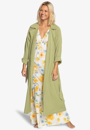 Chaqueta larga ligera de color verde oliva con una tela texturizada, llevada sobre un vestido maxi floral con flores amarillas y azules.