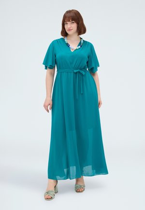 Femme aux cheveux courts et bruns portant une robe maxi turquoise à manches volantes, taille nouée, collier de perles et talons ouverts vert clair, debout sur fond blanc.