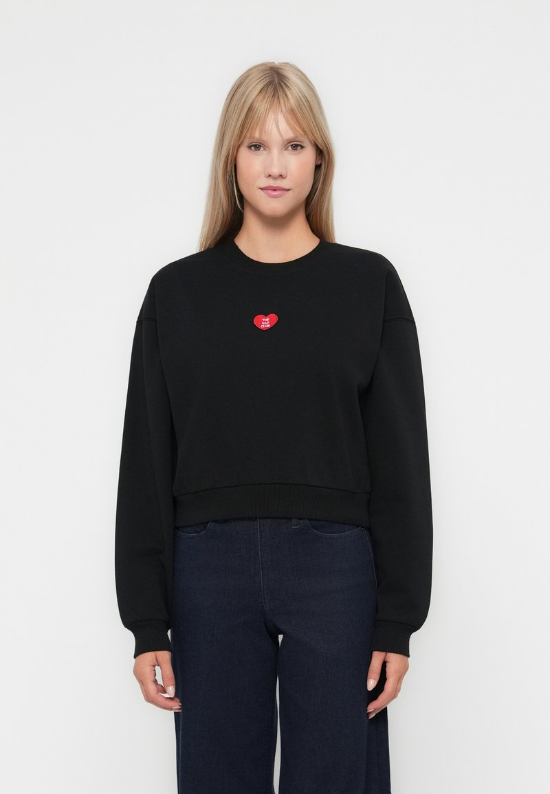 Sweatshirt negru, scurt, cu mâneci lungi și manșete cu dungi, având o aplicație în formă de inimă roșie pe piept. Textură netedă, croi lejer.