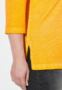 Primo piano della mano di una persona al suo fianco, che indossa una maglietta a maniche lunghe arancione brillante e jeans scuri con piccole borchie metalliche.