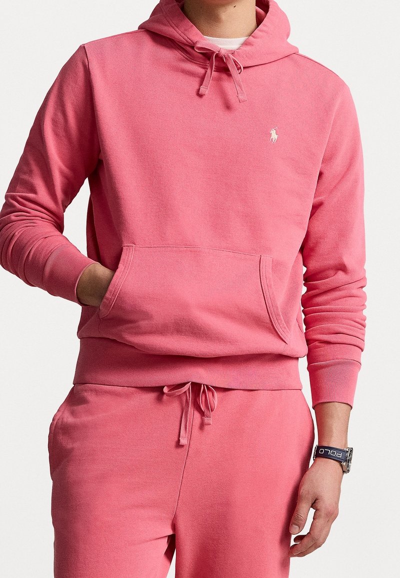 Homme portant un sweat à capuche rose et un pantalon de jogging assorti, la main gauche dans la poche du sweat et une montre au poignet droit.