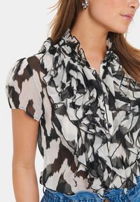 Black and white ruffled blouse made of sheer fabric, featuring a high collar, short sleeves, and decorative button placket details.