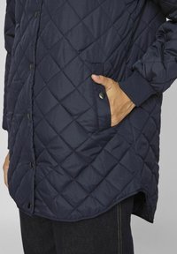 Granatowa pikowana kurtka puchowa z zatrzaskami, prążkowanymi mankietami i boczną kieszenią. Miękki, teksturowany materiał z prostym wykończeniem na dole.